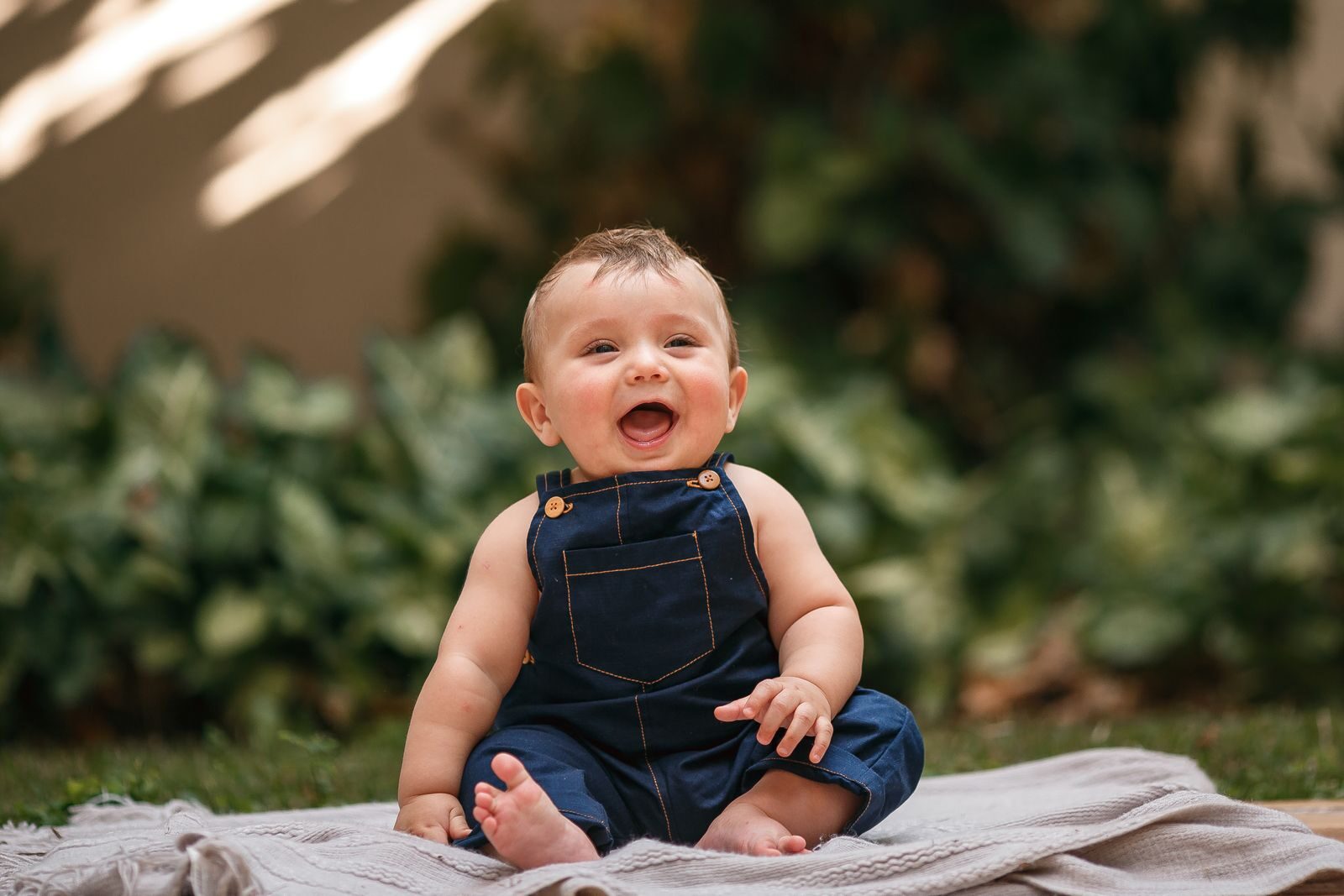 Happy baby in overalls sitting on a blanket outdoors in a garden during summer.